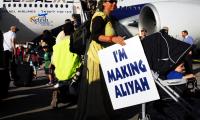 En kvinde ude foran et EL-AL fly med et skilt hvorpå der står 'I'm making Aliyah'